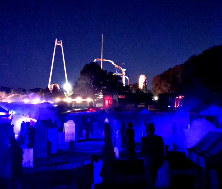 Dark haunted graveyard with glowing blue fog and amusement park rides behind