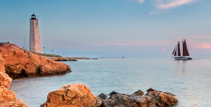 Lighthouse on rocky coast with sailboat on calm sea at sunset