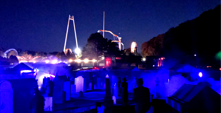 Dark haunted graveyard with glowing blue fog and amusement park rides behind