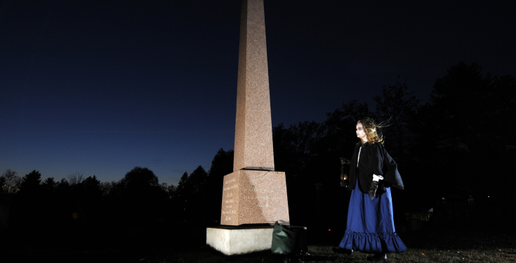 Person holding lantern near tall grave monument at night